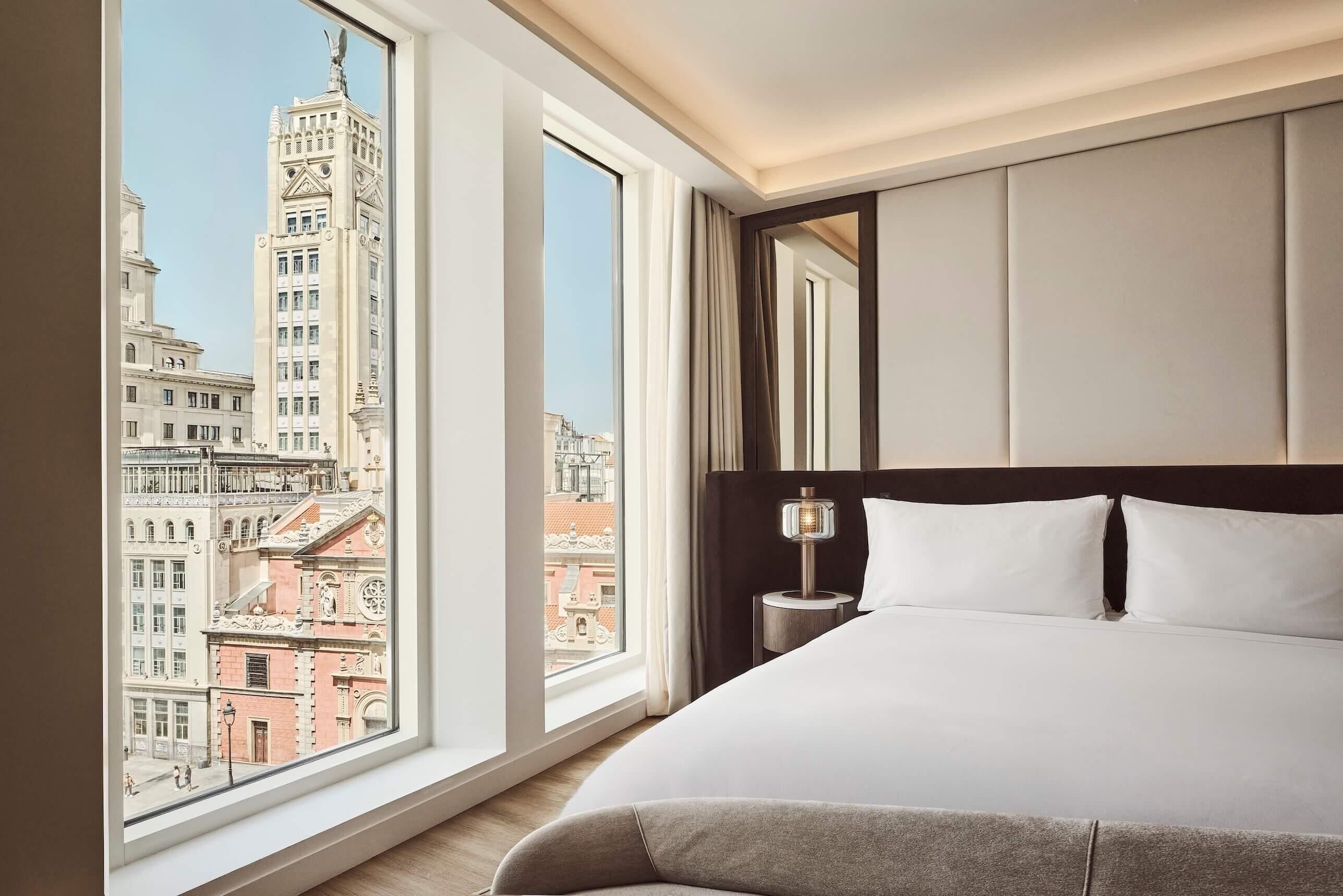 A hotel room with a large bed, a nightstand, and a view of city buildings through large windows.