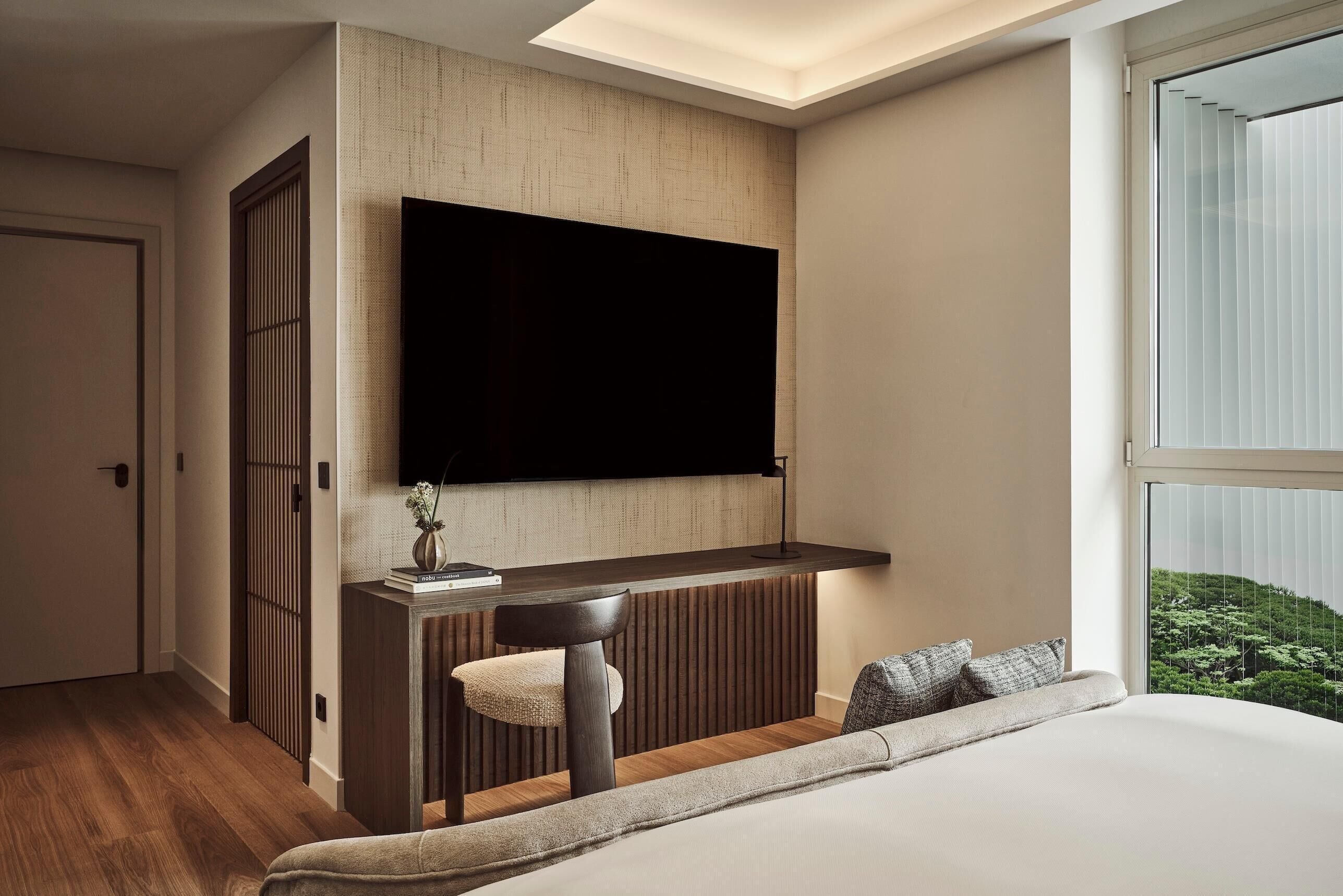 A modern bedroom with a large bed, a mounted TV, a small desk with a chair, and a window with a view of greenery.