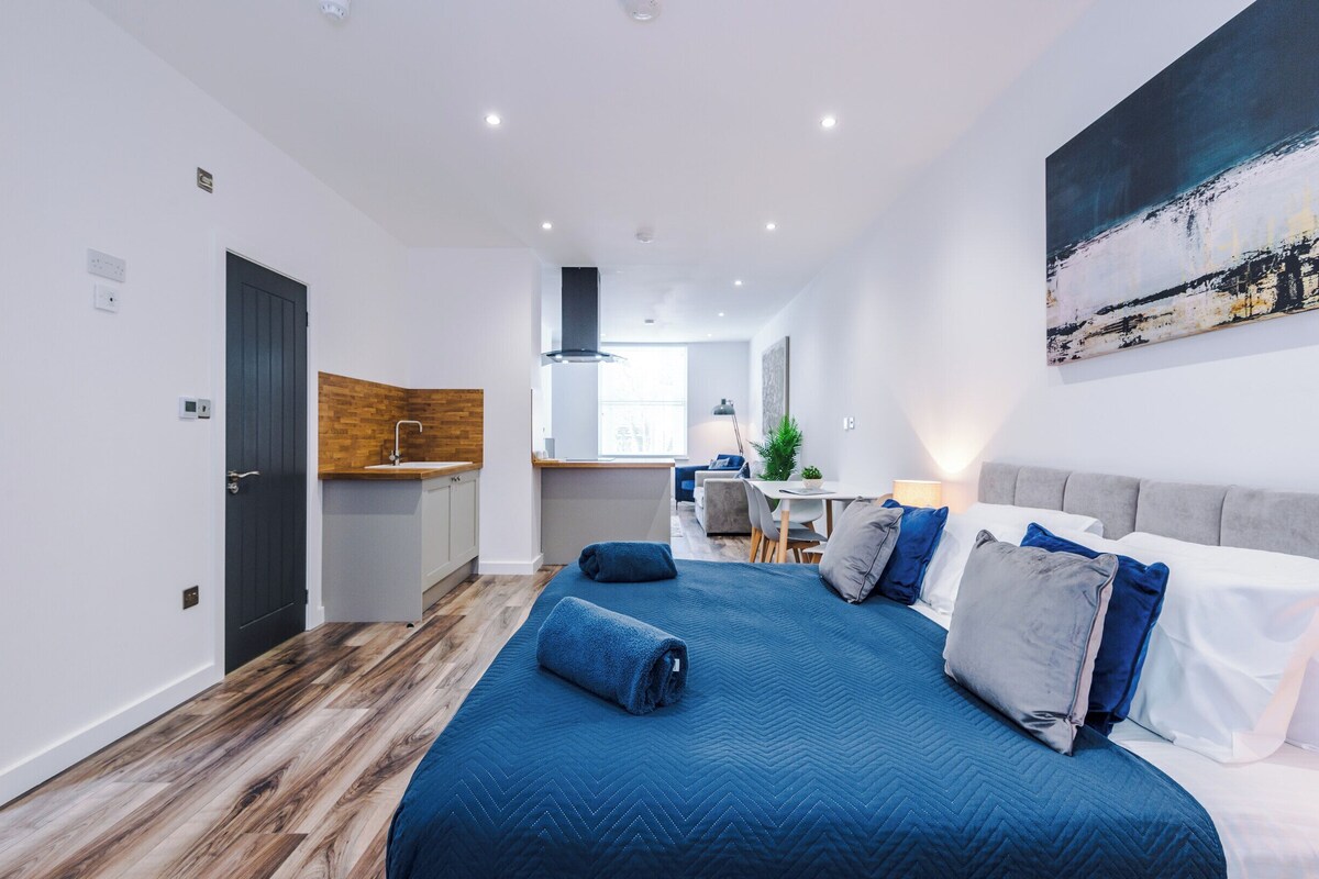 A modern hotel room with a large bed, a desk, a kitchenette, and a view of the city.