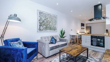 A modern living room with a grey sofa, a blue armchair, a wooden coffee table, and a floor lamp. There is a kitchen area with a stove, oven, and a dining table with chairs. A large abstract painting is on the wall, and a potted plant is visible.