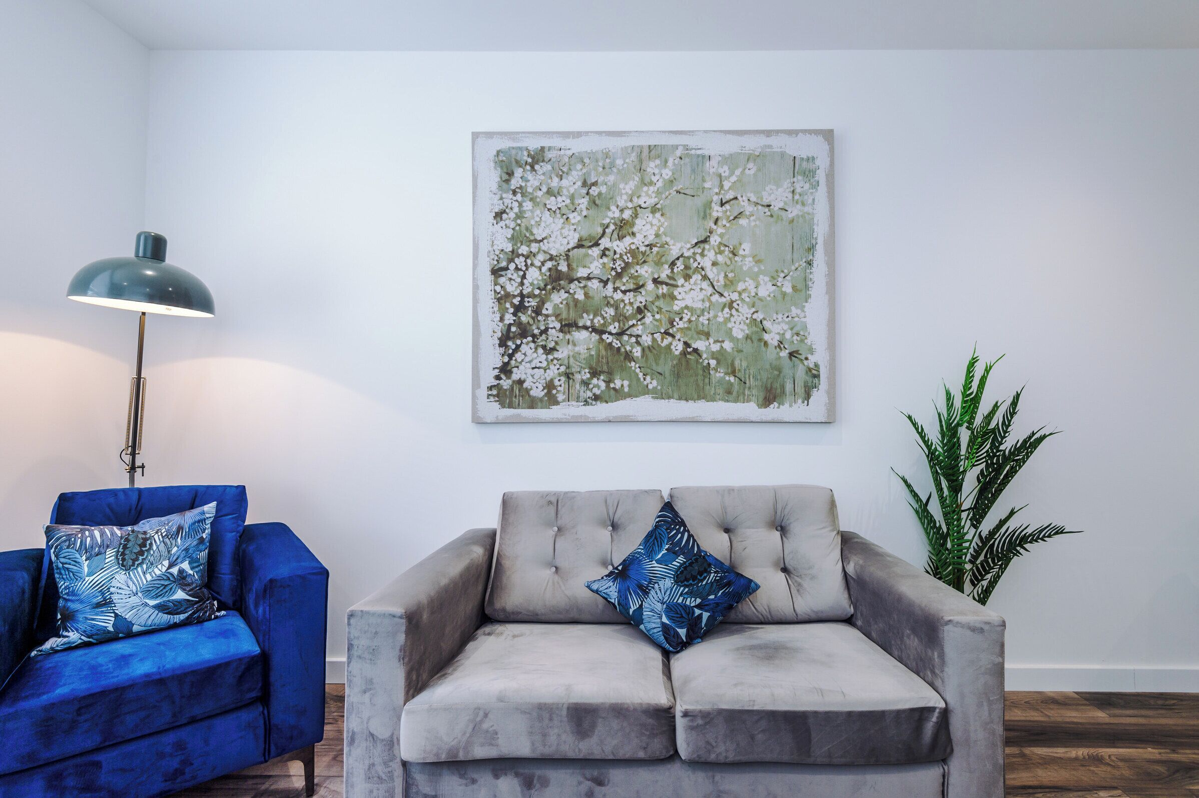 Una sala de estar con un sillón azul, un sofá gris, una lámpara de pie, un cuadro y una planta en maceta.