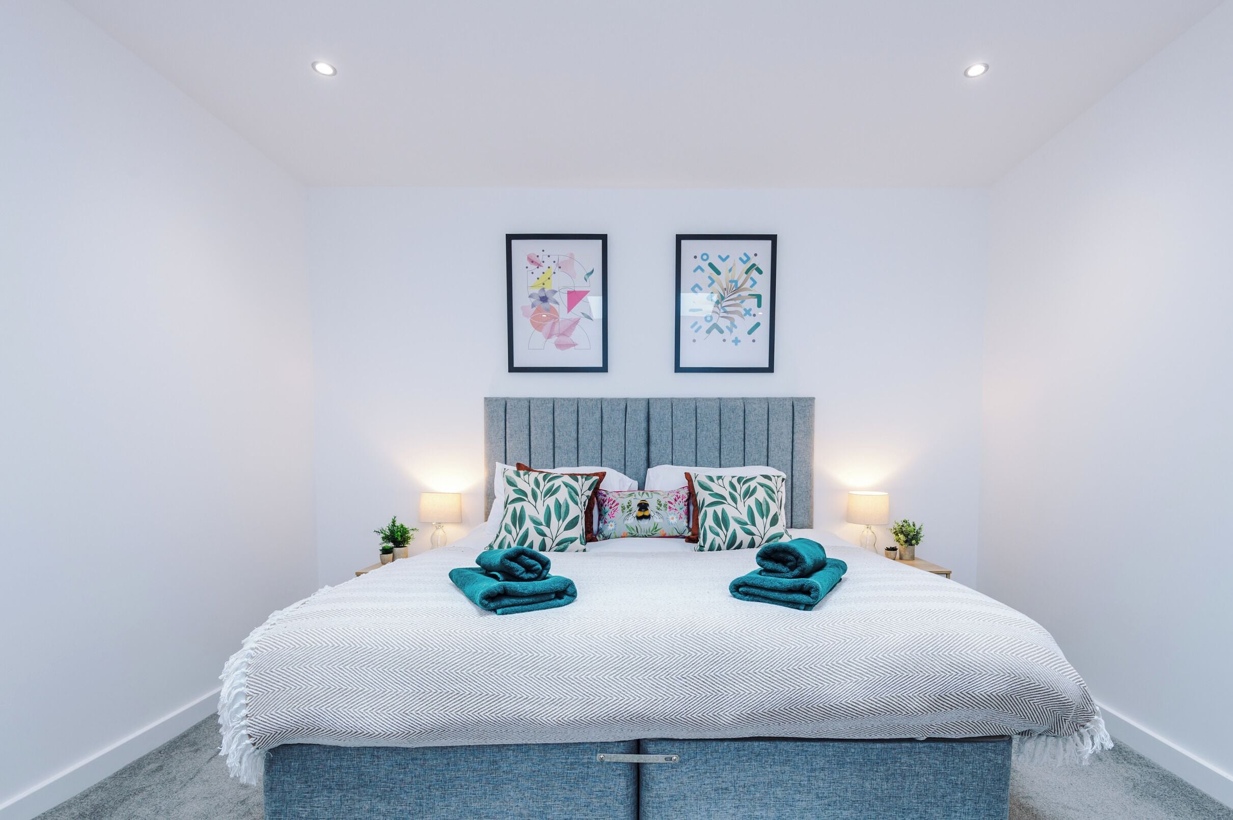 A neatly arranged bedroom with a bed, pillows, two wall-mounted lights, and two framed artworks.