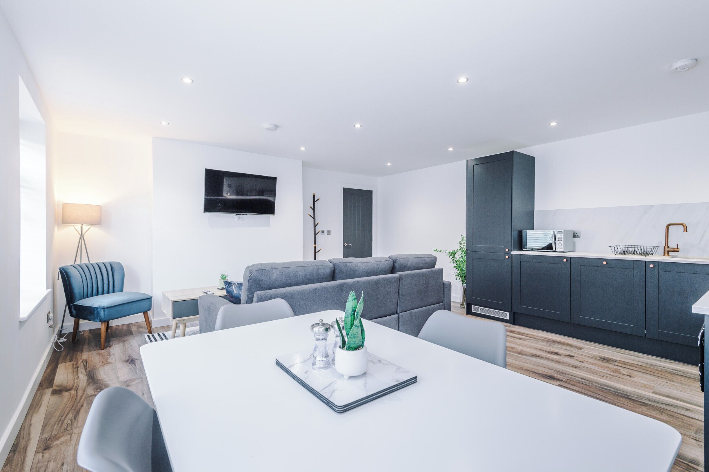 A modern living space with a dining area, grey sofa, kitchen, and a flat-screen TV mounted on the wall.