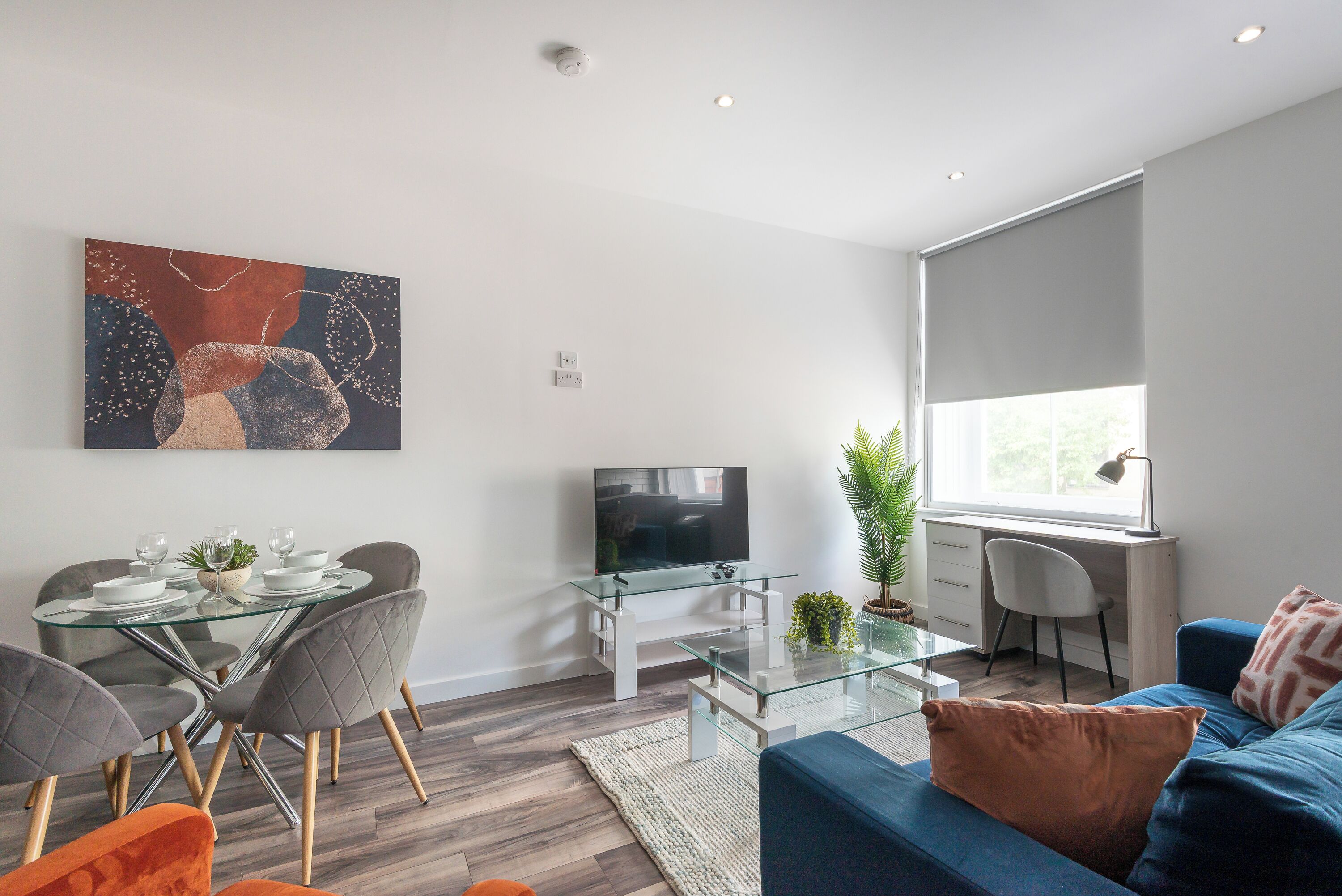 Un salon moderne avec une table à manger en verre, des chaises recouvertes d’un tissu gris, un canapé bleu, un téléviseur à écran plat et un grand tableau abstrait accroché au mur.