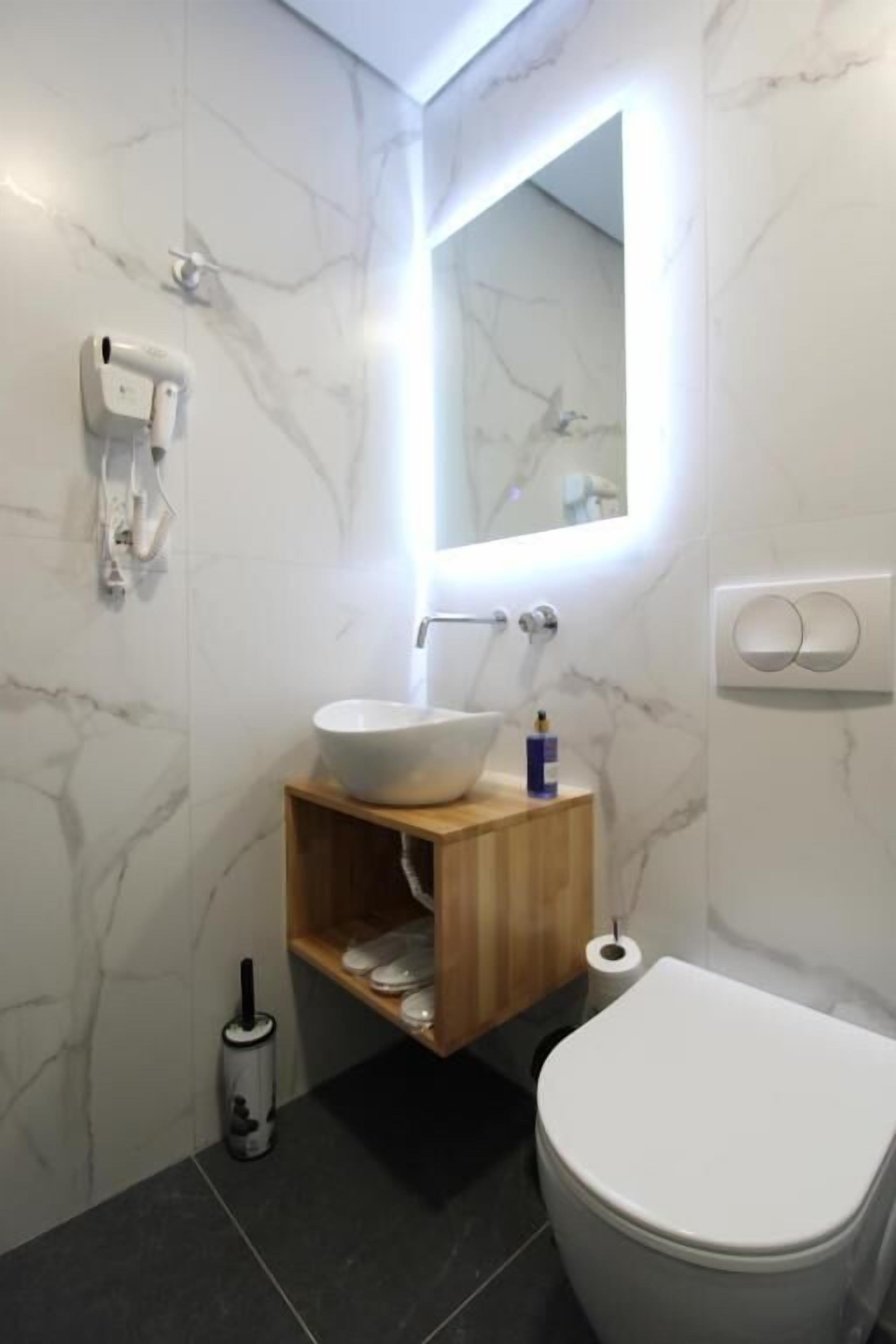 A modern bathroom with a marble wall, a white sink, a wooden vanity, a toilet, and a mirror with a light.