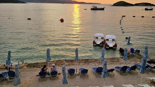 Una playa con sillas y sombrillas, barcos en el agua y una puesta de sol al fondo.