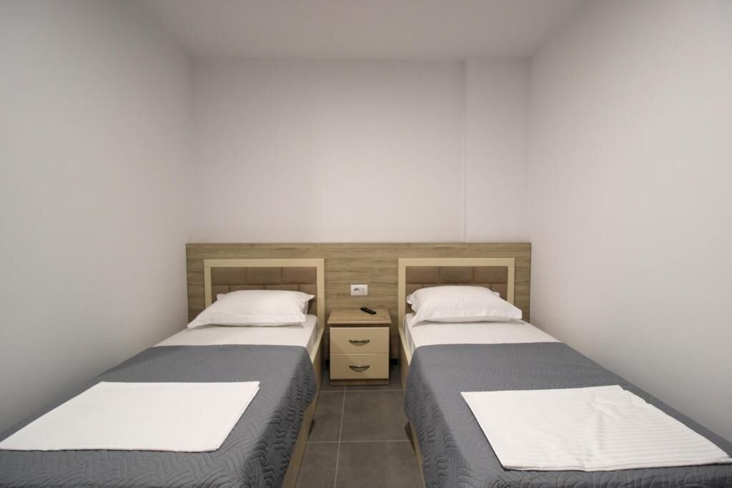 A twin room with two single beds, a wooden headboard, a small bedside table with drawers, and a plain white wall.