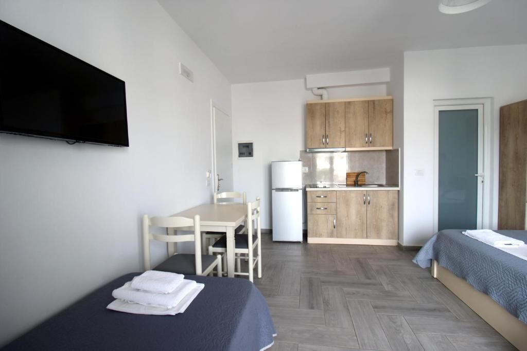 Ein kleines Zimmer mit einem Bett, einem Schreibtisch, einem Stuhl, einem Fernseher und einer Küchenzeile.