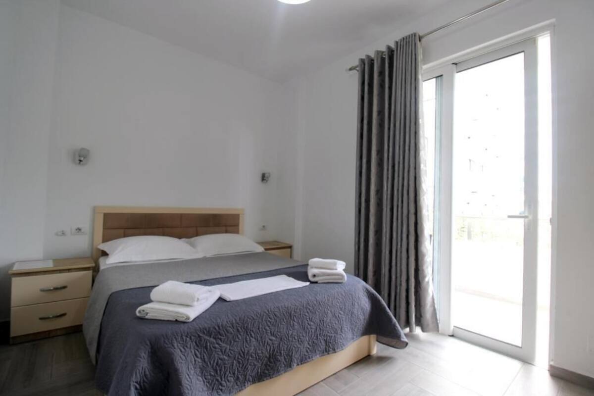 A bedroom with a bed, a nightstand, a window with curtains, and a view of the outside.