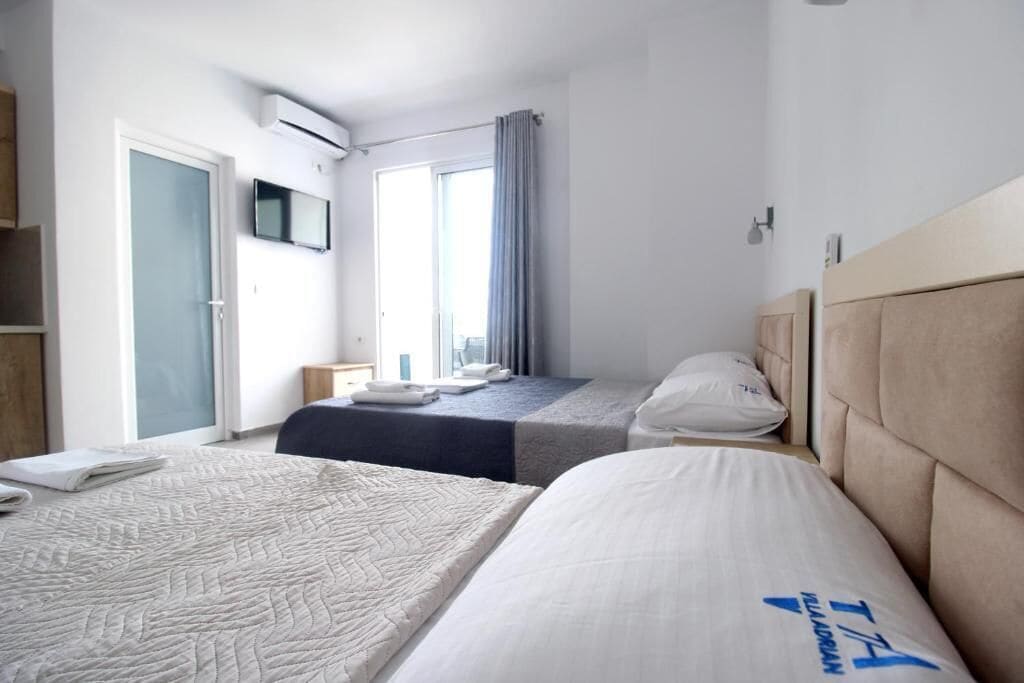 A hotel room with two beds, a TV, an air conditioning unit, a window with curtains, and a view of the city.