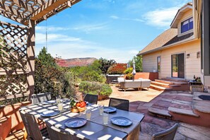 Een terras buiten met een tafel voor vier personen, omringd door stoelen, een houten pergola en uitzicht op een huis en bergen.