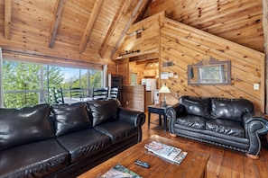 A wooden cabin interior with two leather sofas, a wooden coffee table, and a large window offering a view of greenery. - Private Hot Tub + Deck + Fire Pit + Mountain Views | Mins to Downtown (Townsend)