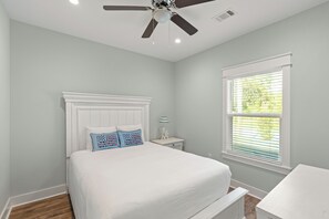 Une chambre à coucher comprenant un lit, un ventilateur de plafond, une table de chevet, une lampe et une fenêtre avec des stores.