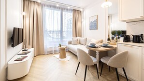 Ein modernes Wohnzimmer mit einer Couch, einem Esstisch, Stühlen, einem Flachbildfernseher und einer Küchenzeile inklusive Kaffeemaschine und Pflanze.
