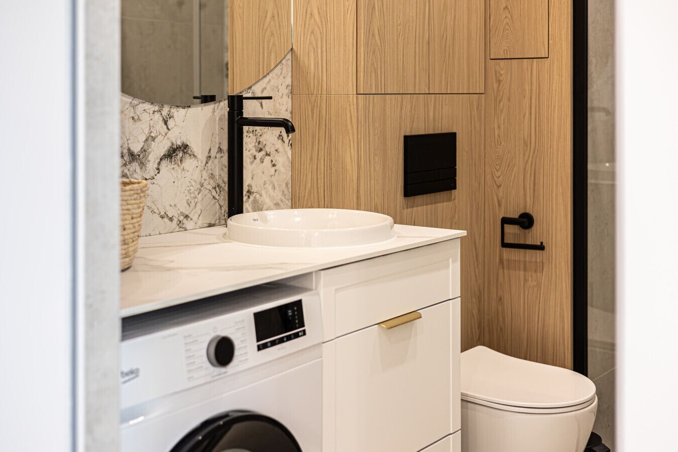A modern bathroom with a built-in washing machine, a sink with a marble backsplash, and a toilet.