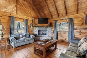 Ein Interieur einer Blockhütte mit Kamin aus Stein, Holzwänden, einer Couch, einem Couchtisch und einem an der Decke befestigten Fernseher.