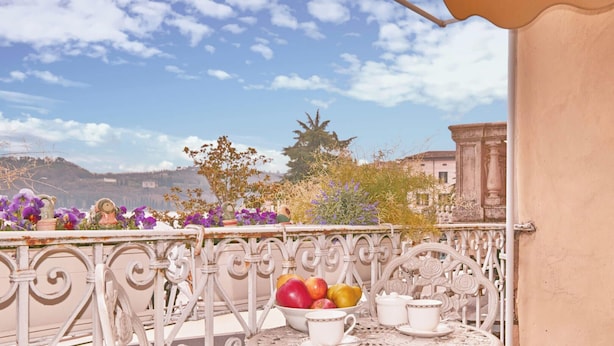 Un balcón con vistas a un paisaje urbano, una mesa con un frutero y una barandilla blanca.