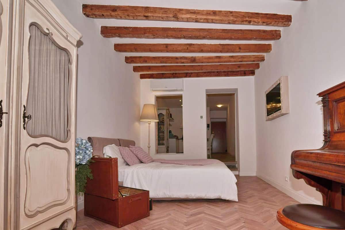 A bedroom with a bed, wooden beams, a dresser, a piano, and a mirror.