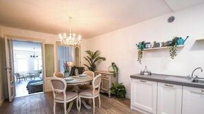 A dining area with a chandelier, a table with chairs, a kitchen with a sink, and a plant shelf.