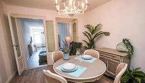 A dining area with a round table, chairs, and a chandelier. There is a plant on the table and a view into another room.