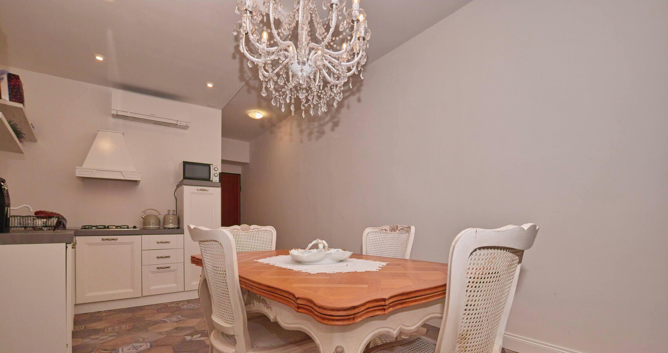 Un'area pranzo con un tavolo in legno, sedie bianche, un lampadario e una cucina sullo sfondo.