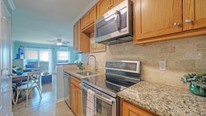 A kitchen with wooden cabinets, stainless steel appliances, granite countertops, and a dining area with a table and chairs. - Walk to Beach + Boardwalk | Golf Cart Zone + Pool Access | Ocean View Efficiency (Port Aransas)
