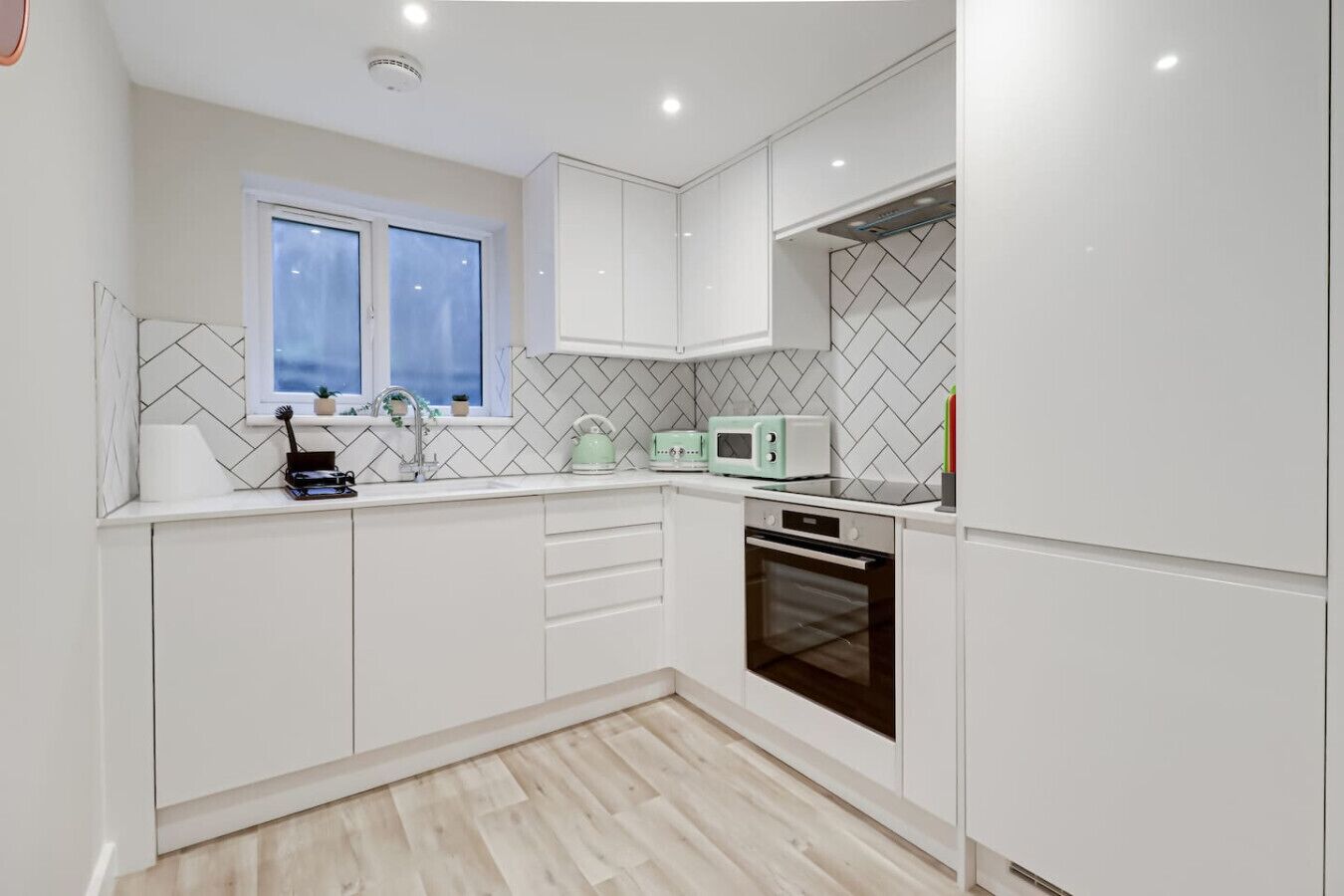 Una cocina moderna con armarios blancos, una ventana, un microondas, un hervidor y un horno.