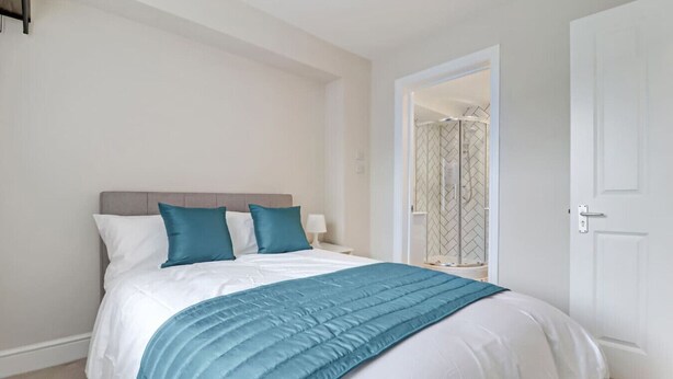 A neatly made bed with a white and teal color scheme, a bedside table with a lamp, and a door leading to a bathroom with a shower.