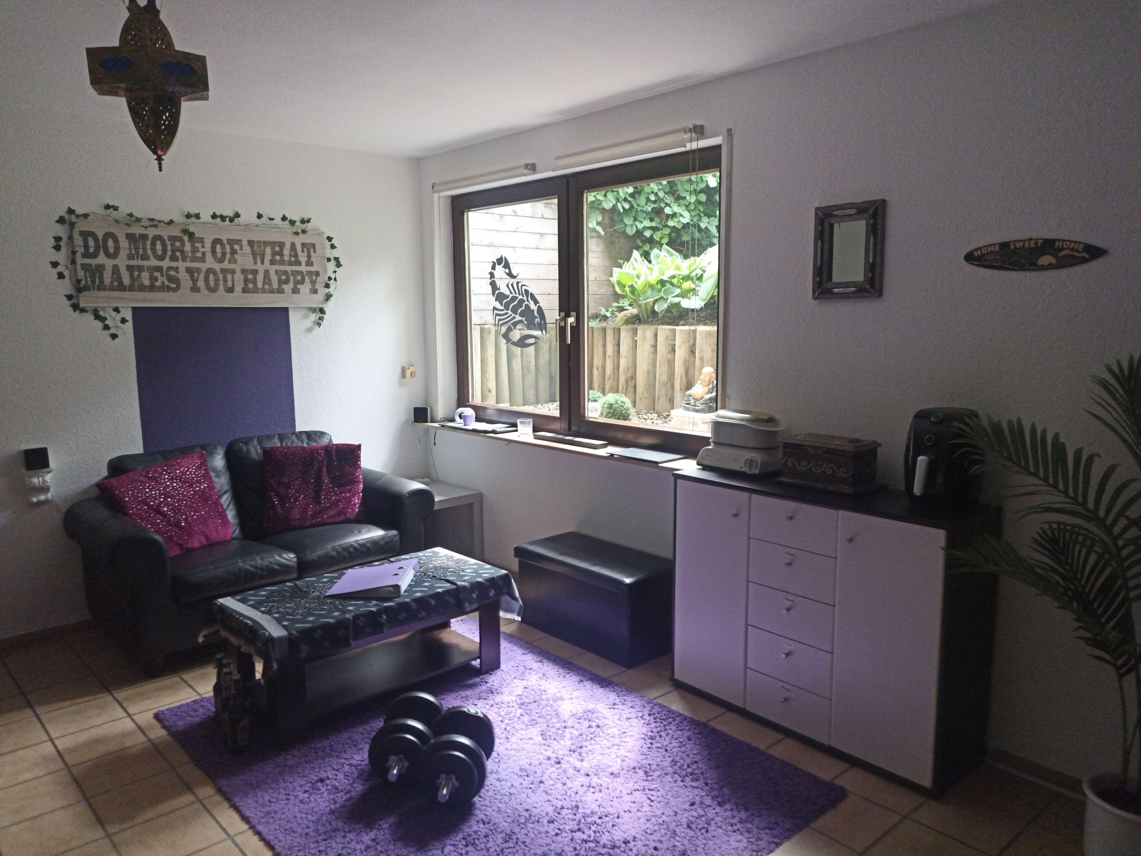 Un espace de vie compact comprenant un canapé, une petite table, une fenêtre donnant sur un espace verdoyant, un meuble et un panneau décoratif accroché au mur.