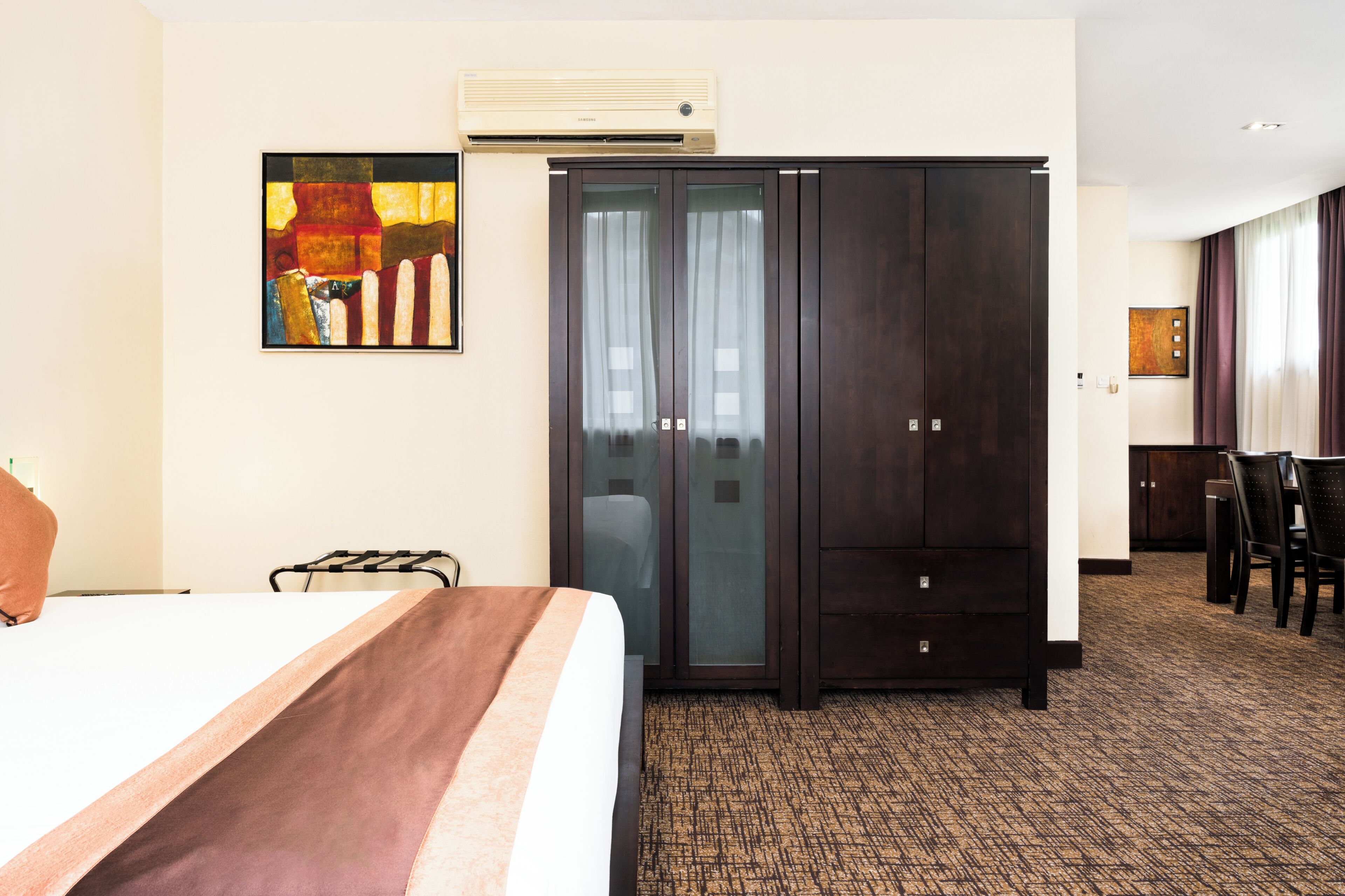 A hotel room with a bed, a wardrobe, a desk with chairs, and a painting on the wall.