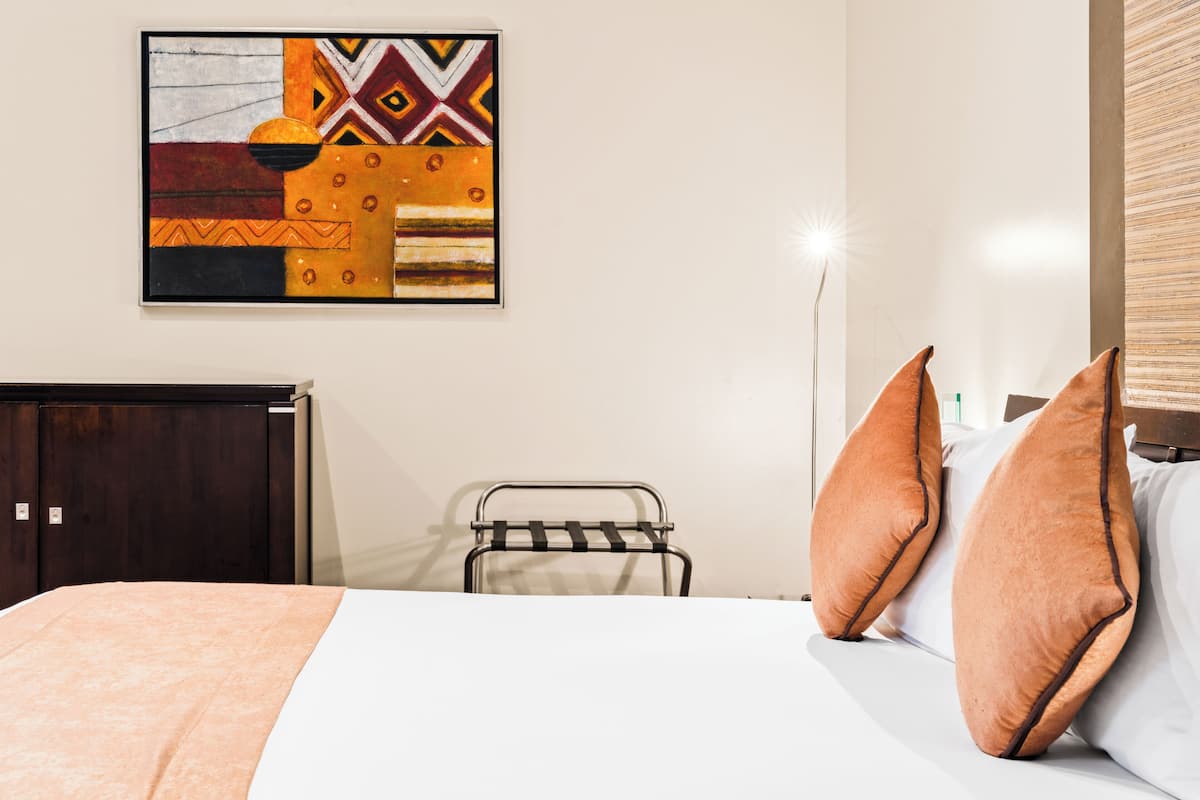 Una habitación de hotel con cama, almohadas, una silla de metal, una obra de arte en la pared y un armario.