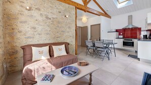 A cozy room with a stone wall, a brown sofa, a dining table with chairs, and a kitchen area with red cabinets. - Gîte Du Côteau – 10 min from Puy du Fou (Les Herbiers)