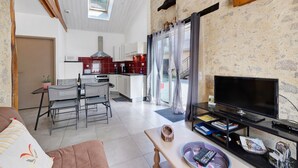 A modern kitchen with white cabinets, a dining area with a table and chairs, a living room with a TV, and a stone wall. - Gîte Du Côteau – 10 min from Puy du Fou (Les Herbiers)
