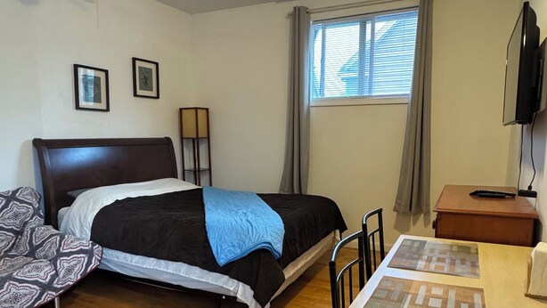 Een slaapkamer met een bed, een bureau, een stoel, een plafondventilator, een raam met gordijnen en een aan de muur bevestigde televisie.