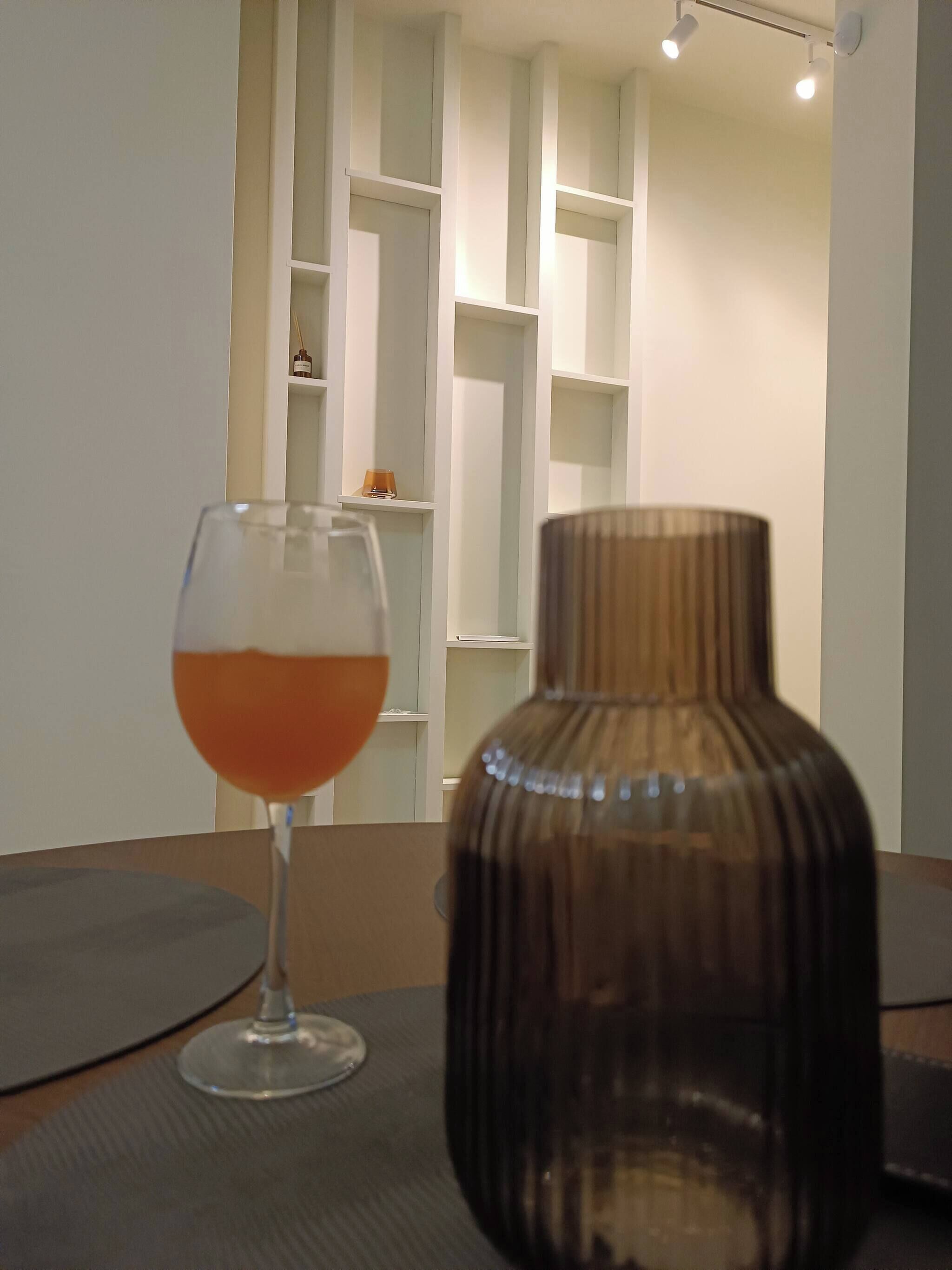 Un verre contenant un liquide orange est posé sur un tapis, à côté d’un vase foncé et d’une étagère blanche, dans une pièce.