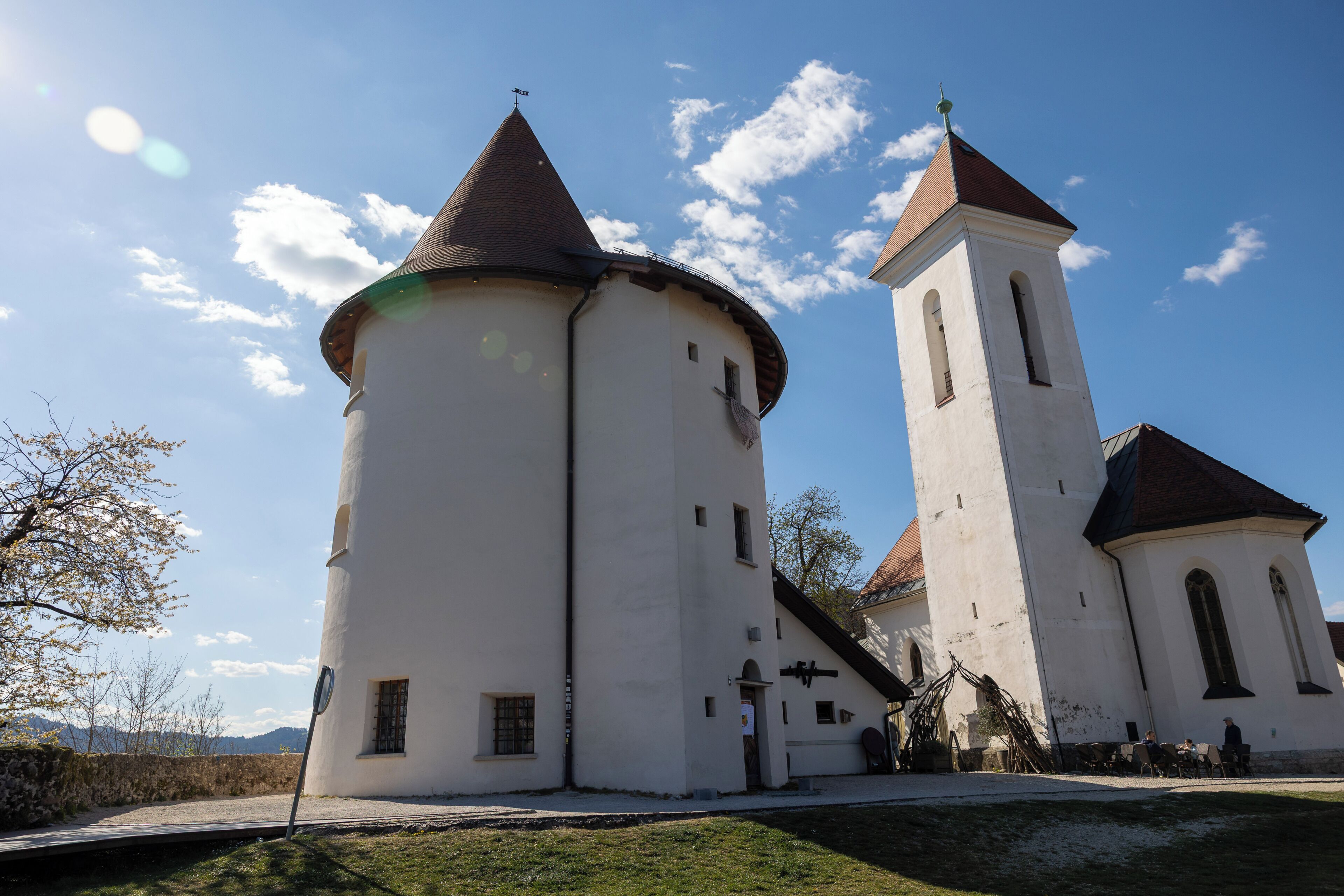 A church with a bell tower and a round tower, surrounded by a grassy area.