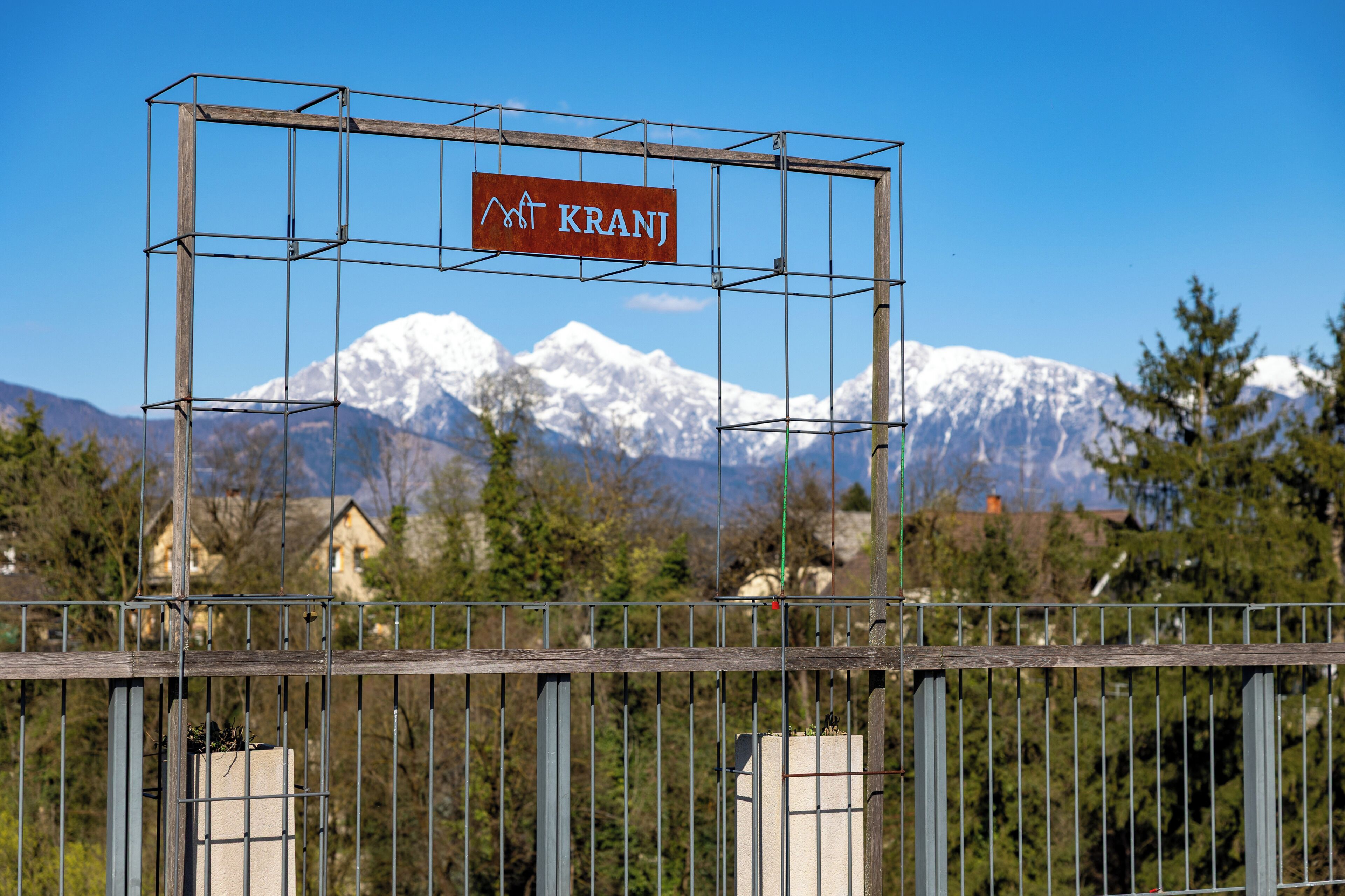 A metal structure with a sign that reads 'Kranj' against a backdrop of snow-capped mountains and houses.