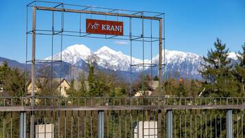 A metal structure with a sign that reads 'Kranj' against a backdrop of snow-capped mountains and houses.