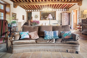 Ein gemütliches Wohnzimmer mit braunem Sofa, gemusterten Kissen, Holzbalken und einer Kristallleuchter.