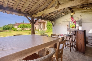 A rustic outdoor dining area with a wooden table and wicker chairs, a stone floor, and a view of houses in the background. - Guest Room - Les Jardins de la Beylie (Château-l'Évêque)