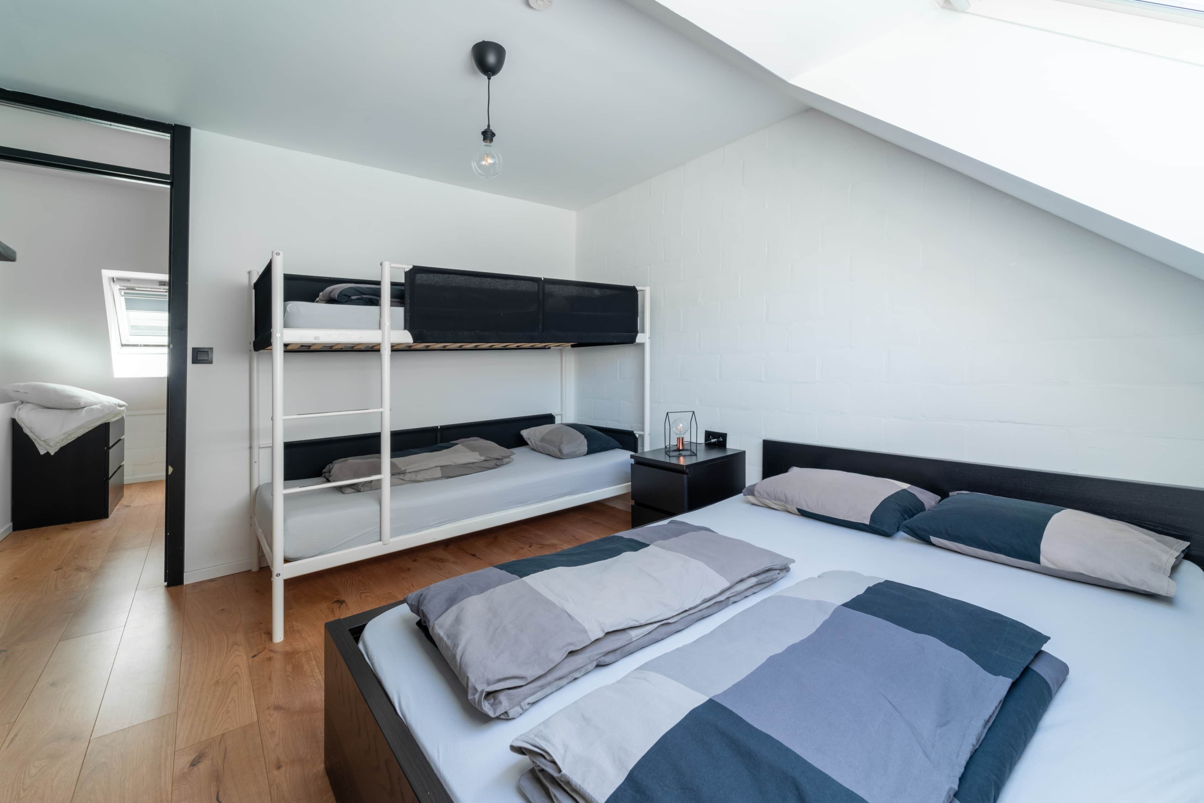 A modern bedroom with a bunk bed, a single bed, a bedside table with a lamp, and a window providing natural light.