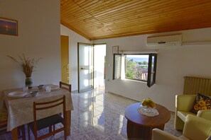 A dining area with a table set for four, a sofa, a window with a view, and a ceiling fan. - Shared gardened accommodation with bbq and sea view (Zambratija)