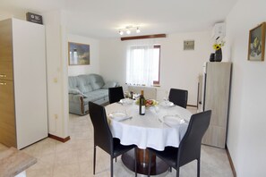 Ein Essbereich mit einem runden Tisch, gedeckt für vier Personen, umgeben von schwarzen Stühlen. Links befindet sich ein Kühlschrank, ein Fenster mit Vorhängen und ein Bild an der Wand.