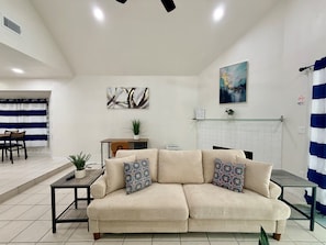 A living room with a beige sofa, a dining table, and chairs. There is a fireplace, a painting, and a plant on the wall. - Waterside at VICC - Cozy, Blissful Views! (Brownsville)