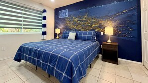 A bedroom with a bed, a ceiling fan, a nightstand, a lamp, a window with blinds, and a wall with a cityscape mural. - Waterside at VICC - Cozy, Blissful Views! (Brownsville)