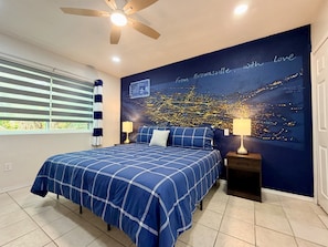 A bedroom with a bed, a ceiling fan, a nightstand, a lamp, a window with blinds, and a wall with a cityscape mural. - Waterside at VICC - Cozy, Blissful Views! (Brownsville)