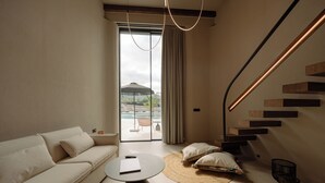 Una sala moderna con un sofá, una mesa de centro redonda, una escalera de madera y vistas a un área exterior con piscina y sombrillas.