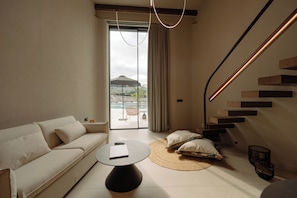 A modern living room with a sofa, a round coffee table, a staircase with wooden steps, and a view of an outdoor area with a pool and umbrellas. - Superior Villa - Kyklio Pavilions Nafplio (Nafplio)