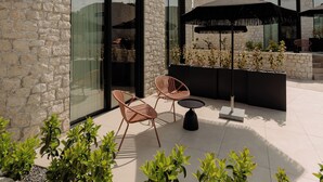 Un edificio de piedra con grandes ventanales, asientos exteriores con sillas y una mesa, y un paraguas negro.