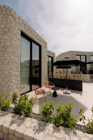 A stone building with large glass windows, outdoor seating with chairs and a table, and a black umbrella. - Superior Villa - Kyklio Pavilions Nafplio (Nafplio)